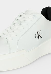 Baskets basses blanches avec lacets blancs, semelle épaisse, doublure intérieure noire et logo "CK Calvin Klein" noir sur le côté.