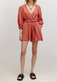 Koraal kleurige jumpsuit in wikkelstijl met pofmouwen, een strik taille, textuurstof en een diepe V-hals, gecombineerd met zwarte slidesandalen.