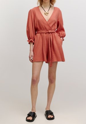 Coral wrap-style playsuit with puff sleeves, a tie waist, textured fabric, and a deep V-neck, paired with black slide sandals.