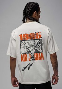 T-Shirt mit kurzen Ärmeln aus cremefarbenem Baumwollstoff, das ein großes schwarzes und oranges Grafikmotiv eines Basketballspielers sowie den Text "1985 AIR JORDAN" auf der Rückseite zeigt.