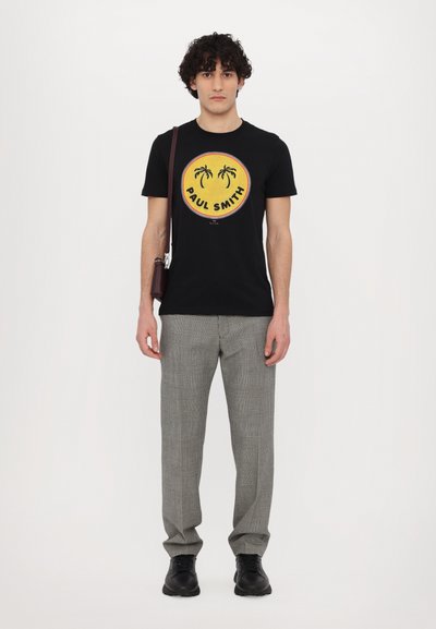 T-shirt noire avec un logo circulaire jaune représentant des palmiers et le texte "PAUL SMITH", associée à un pantalon à carreaux gris et des chaussures noires.