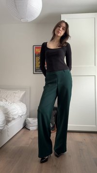 Mujer de pie en interior, vistiendo una blusa negra de manga larga, pantalones anchos verde oscuro, tacones negros, con una cama y un póster enmarcado al fondo.