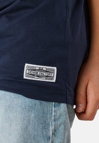 Laivasilmäinen puuvillapaita, jossa on valkoinen kudottu etiketti, jossa lukee "Original Denim Factory." Paidassa on huolitellut reunat; kangas näyttää sileältä.