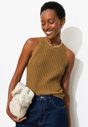 Femme souriante portant un haut marron sans manches en maille et des bijoux en or, tenant un sac en tissu clair à motifs, debout devant un fond blanc.