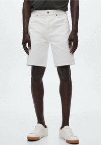 Persona vestida con pantalones cortos de mezclilla blancos, camiseta negra y zapatillas blancas con suelas de goma, de pie contra un fondo liso claro.