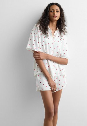 Frau mit lockigem Haar, die ein weißes, kurzärmliges Hemd mit Knöpfen und passende Shorts mit kleinen roten Erdbeeraufdrucken trägt.