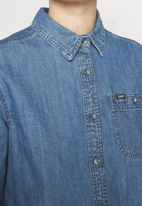 Jeansskjorta i medelblått med knappslå, spetsig krage och en enda bröstficka med en liten märkesetikett. Strukturerad tyg.