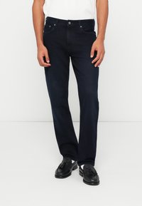 Tamsiai mėlynos džinsinės kelnės su tiesiu siluetu, su priekiniais mygtukais, penkių kišenių dizainu ir šiek tiek nudilusiu paviršiumi.