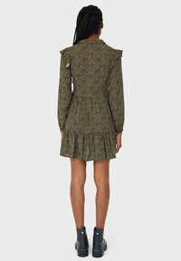 Vihreä kuvioitu mekko, jossa on röyhelöiset olkapäät, kapea vyötärö ja kerroksellinen hame. Yhdistetty mustiin nilkkabootseihin.