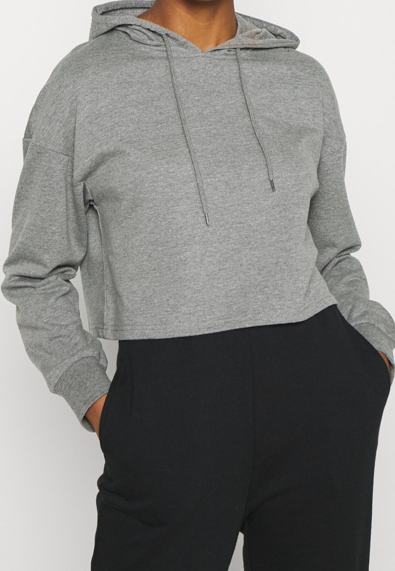 Personne portant un sweat à capuche gris court avec cordons et un pantalon noir taille haute, les mains dans les poches, sur un fond uni.