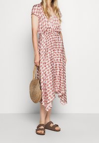 Maxi-jurk in het wit met een rood bladpatroon, korte mouwen, elastische taille, vloeiende vorm. Geweven stro tas en bruine sandalen complementeren de outfit.