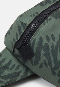 Zbliżenie na zielony worek z tkaniny w kamuflażu z czarnym suwakiem i kwadratowym uchwytem, z wytłoczonym półokrągłym logo.