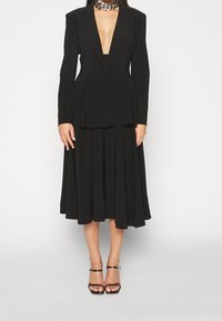 Černý blazer s hlubokým výstřihem do V a strukturovanými rameny, v kombinaci s volnou černou midi sukni. Hladká textura a minimální kovové akcenty.