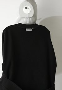 Felpa nera con scollo rotondo e polsini a coste. Presenta un motivo geometrico testurizzato sul retro. Il tessuto sembra morbido e spesso.