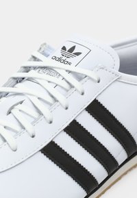 Bijele kožne tenisice s tri crne horizontalne pruge, bijelim vezicama i oznakom Adidas na jeziku. Teksturalna gumena potplat.