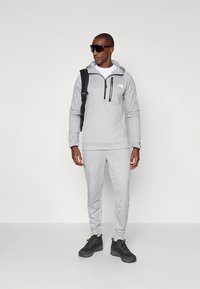 Sweatshirt e calças de fato de treino cinzentos com um bolso no peito com fecho, usados com óculos de sol pretos e uma mochila, apresentando um tecido texturizado e um corte descontraído.