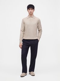 Homme debout portant un pull polo beige à manches longues, un pantalon bleu marine foncé et des mocassins beiges sur un fond blanc uni.
