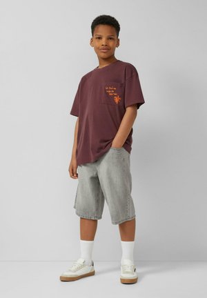 Garçon debout, une main dans la poche, portant un t-shirt bordeaux avec texte sur la poche et un palmier, short gris, chaussettes blanches et baskets sur fond uni.