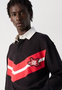 Giovane con capelli ricci scuri che indossa una maglia da rugby nera con colletto bianco e una striscia a V rossa e bianca con stemma sul petto.
