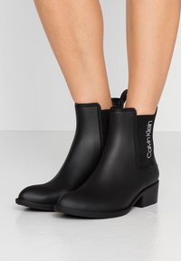 Bottines noires avec une finition lisse et mate. Elles possèdent des panneaux latéraux élastiques et un petit talon cubain. Le logo Calvin Klein est affiché de manière proéminente.