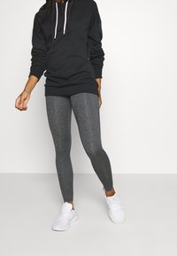 Czarna bluza z kapturem z białymi sznurkami, w zestawieniu z szarymi legginsami z rozciągliwego materiału i białymi butami sportowymi, w pozycji stojącej.