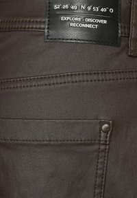 Donkerbruin denim kledingstuk met een leren label met coördinaten en de tekst "VERKEN - ONTDEK - HERSTEL," eenvoudige achterzakontwerp.