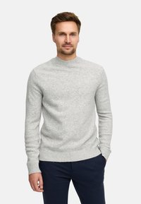 Uomo con capelli corti castani che indossa un maglione lavorato a maglia grigio chiaro e pantaloni blu scuro, mentre è in piedi contro uno sfondo bianco semplice.