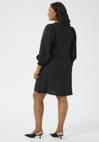 Abito nero con tessuto texturizzato, maniche lunghe a sbuffo e vestibilità ampia, che finisce sopra il ginocchio. Indossato con tacchi a slingback neri.