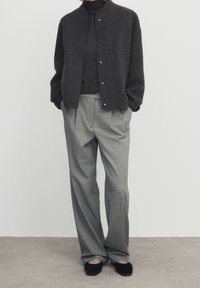 Veste en laine grise foncée à boutons, portée par-dessus un col roulé gris, accompagnée de pantalons gris clair à plis et de chaussures plates noires.