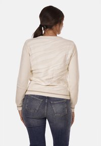 Maglione color crema con un motivo a onde texturizzato, polsini e orlo a costine. Indossato con jeans blu scuro, che mostrano una silhouette aderente.