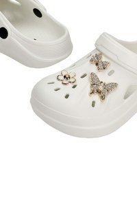 Sabots blancs à enfiler avec trous de ventilation, décorés de charms papillon, nœud et fleur en ton doré sertis de pierres.