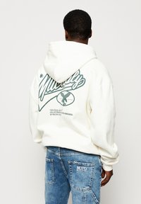Hoodie bianco con stampa grafica verde sul retro, che presenta un testo in corsivo e un design di aquila. Abbinato a jeans blu sbiaditi con toppa del logo.