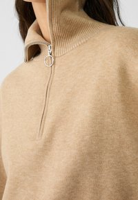 Beżowy sweter z wysokim kołnierzem oraz detalem w postaci zamka błyskawicznego. Wykonany z miękkiego materiału, ma prążkowaną strukturę przy kołnierzu i okrągły uchwyt zamka.