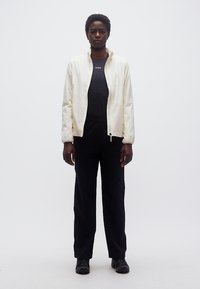 Άτομο στέκεται αντικρίζοντας προς τα εμπρός, φορώντας λευκό μπουφάν με φερμουάρ πάνω από μαύρο πουκάμισο, μαύρο παντελόνι και μαύρα παπούτσια, μπροστά σε απλό λευκό φόντο.