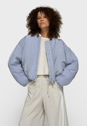 Paskowana niebiesko-biała krótka kurtka bomberka z zamkiem błyskawicznym i oversize'owymi rękawami, zestawiona z jasnoszarymi, wysokim stanem spodniami.
