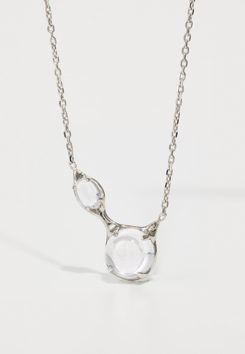 Caurspīdīgs kulons ar kaklarotu, kas ietver divas apaļas stikla pērles, iestiprinātas sudraba tonī metāla rāmi ar saistītu ķēdi.