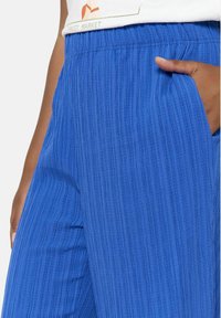 Modre teksturirane hlače z nagubanimi detajli, elastičnim pasom in stranskimi žepi. Material se zdi lahek in zračen.