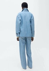 Jasnoniebieska kurtka dżinsowa z wysokim kołnierzem, luźnym krojem i bocznymi kieszeniami, zestawiona z pasującymi spodniami typu wide-leg, pokazana z tyłu.