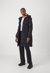Piumino nero con cappuccio, caratterizzato da un design trapuntato, bottoni frontali e tasche laterali. Indossato sopra una camicia bianca e pantaloni neri.