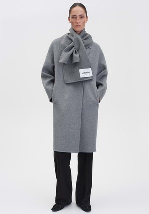 Woman wearing a gray long coat with matching scarf labeled "someday." black pants and black shoes, standing against a plain white background.