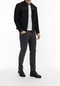 Mann trägt eine schwarze Lederjacke, ein weißes T-Shirt, dunkle graue Jeans und schwarze Schnürstiefel, steht mit einer Hand in der Tasche.