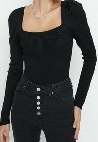 Zwarte geribbelde body met lange mouwen, vierkante halslijn en opgezwollen schouderdetails, gestyled met hooggetailleerde zwarte jeans met metalen knoopaccenten.