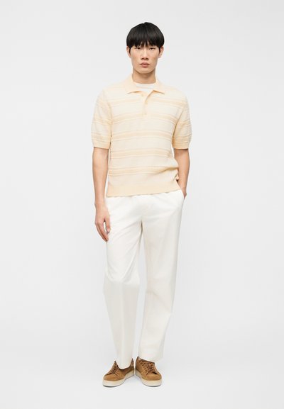 Πόλο με κοντά μανίκια σε ανοιχτό ροδακινί χρώμα με υφασμάτινες ρίγες, συνδυασμένο με λευκό παντελόνι και καφέ αθλητικά παπούτσια. Απλός γιακάς με λεπτομέρεια κουμπιών.