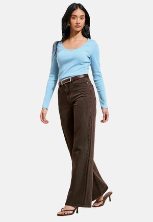 Kvinde med langt mørkt hår iført lyseblå bluse med lange ærmer, brune high-waist viddebukser, brun bælte og brune hælede sandaler, der går fremad.