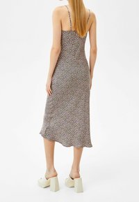 Kobieta z długimi blond włosami, ubrana w bezrękawową, kwiecistą sukienkę midi oraz białe sandały na słupku z otwartym tyłem, stojąca na białym tle.