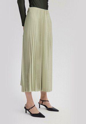 Ženska v svetlo zeleni nagubani midi krilu in črnih čevljih z nizko peto z ostrim koničastim peto na enostavni podlagi.