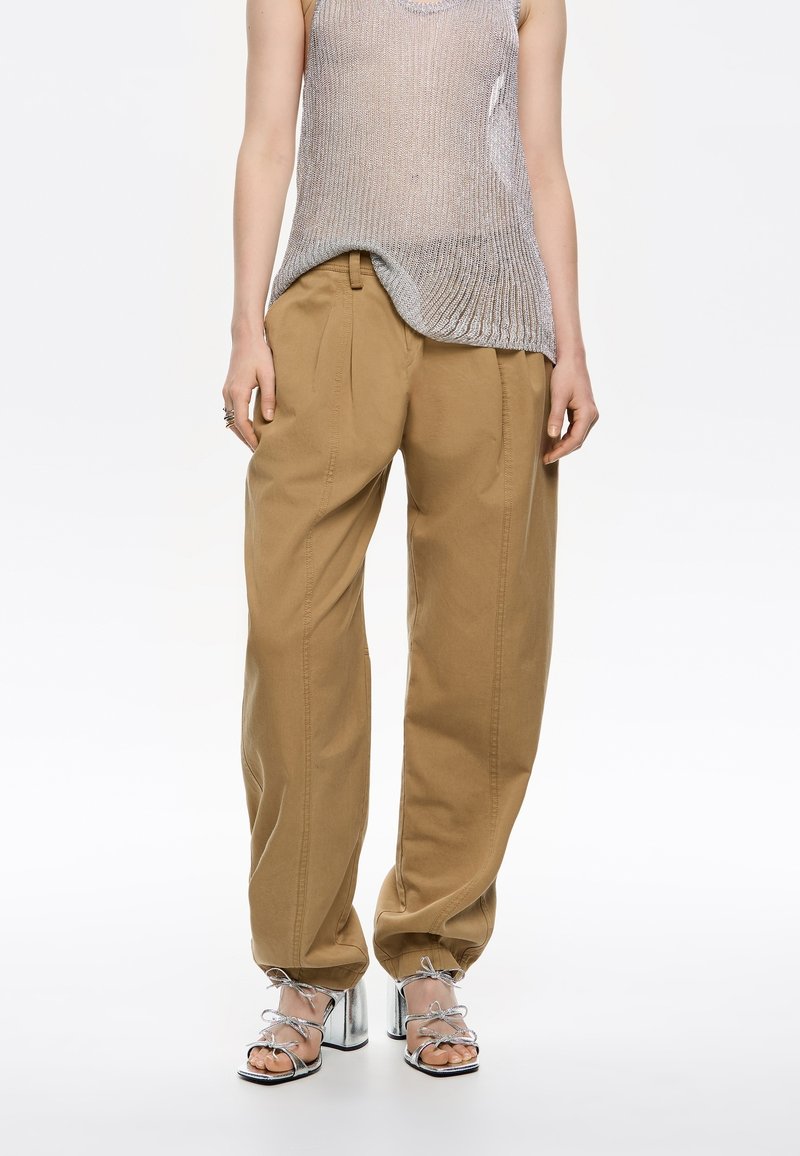 Pantaloni beige a gamba larga in cotone. Il design presenta una vestibilità rilassata con pieghe frontali, abbinati a sandali con tacco a spillo argentati.