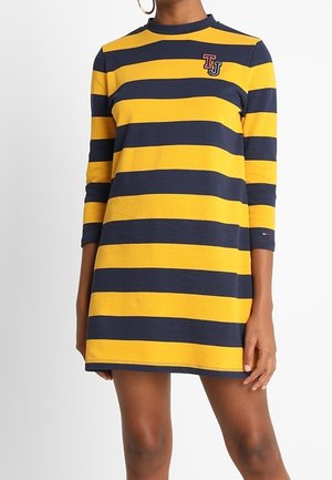 Mujer vistiendo un vestido a rayas horizontales en amarillo y azul marino con mangas tres cuartos y un pequeño logo bordado "TJ" en el pecho.