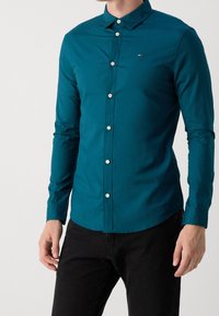 Turkooise overhemd met knopen en lange mouwen, gemaakt van gladde stof. Voorzien van witte knopen en een klein logo op de borst. Gecombineerd met een zwarte broek.