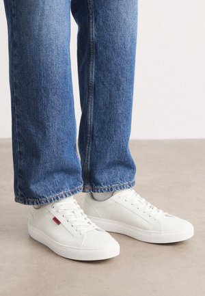 Personne portant un jean bleu et des baskets blanches basses, debout sur un sol clair sur fond uni.